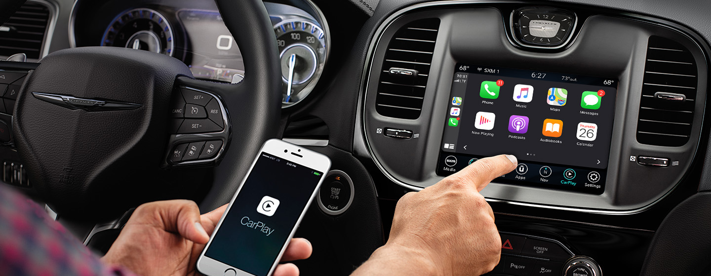 Un conductor sostiene un smartphone y se acerca a la pantalla táctil en el Chrysler 300 2021, donde se ven íconos de conectividad.