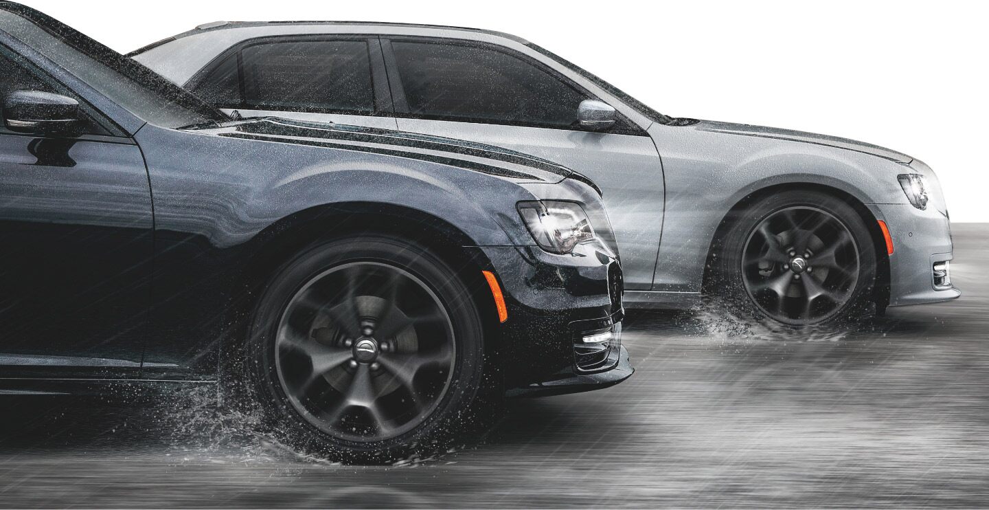 Dos modelos de Chrysler 300 2023 andando en una carretera mojada por la lluvia, con agua que salpica de las ruedas.