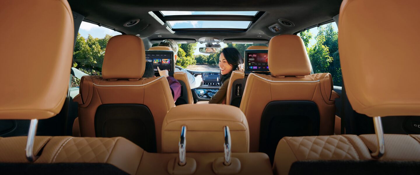 El interior de la Chrysler Pacifica Pinnacle Hybrid 2023 con un hombre conduciendo y una mujer mirando hacia atrás a un niño en la segunda fila que está jugando en la pantalla táctil instalada en el respaldo del asiento.