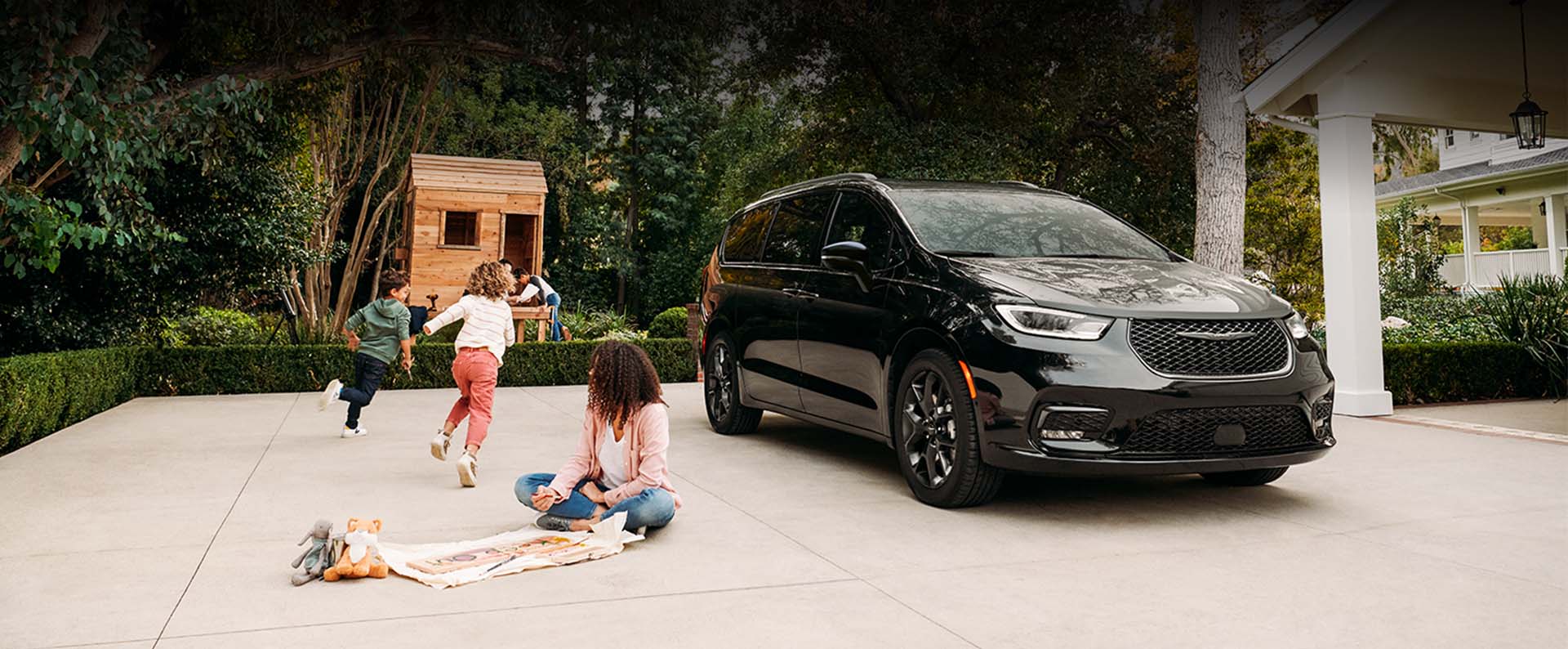 Una Chrysler Pacifica Limited 2024 negra, con el paquete de apariencia S, estacionada en la entrada de una casa residencial con niños jugando a su lado.