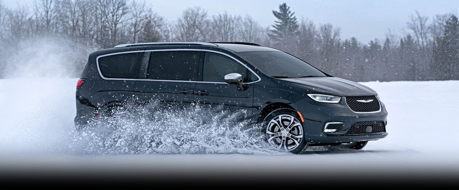 Una Chrysler Pacifica Pinnacle 2025 negra circulando por un camino cubierto de nieve en el bosque.