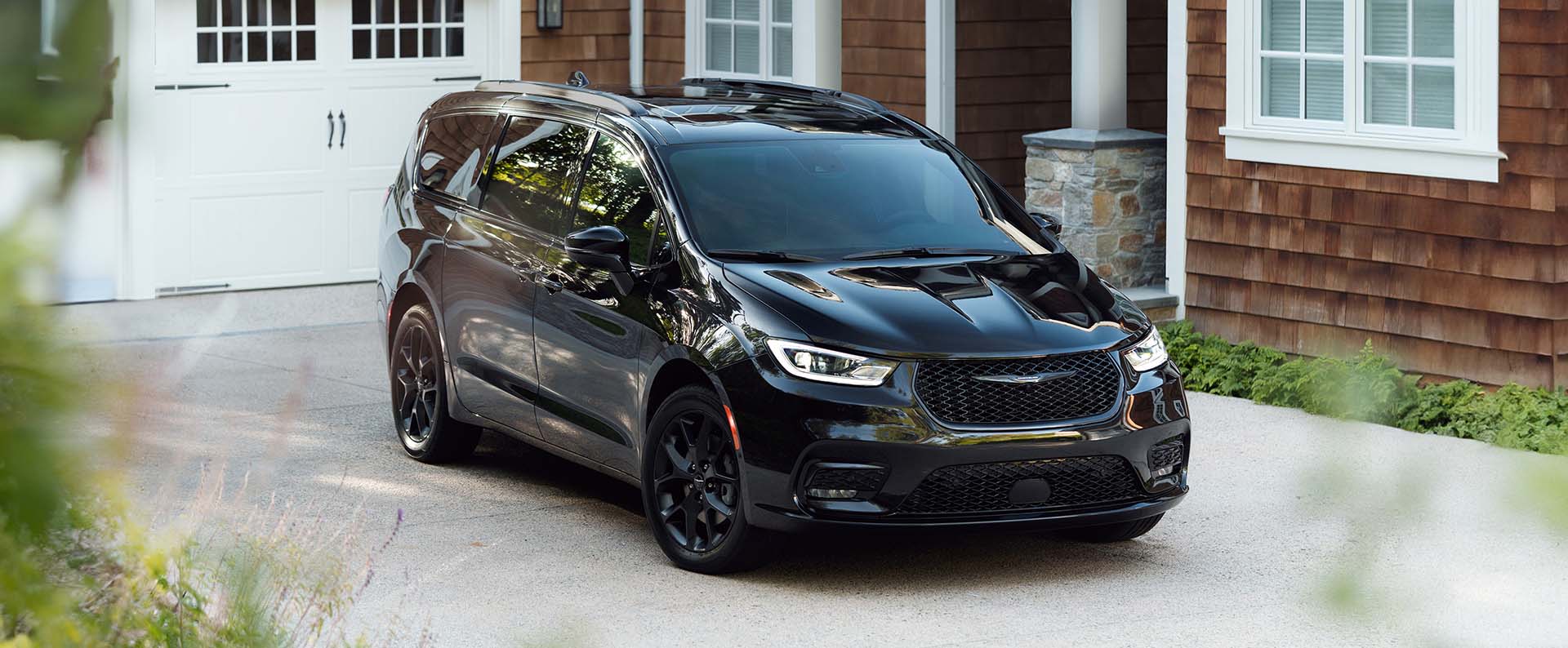 Ángulo delantero del lado del pasajero de una Chrysler Pacifica Limited 2025 negra, estacionada en la entrada de garaje de una casa moderna.