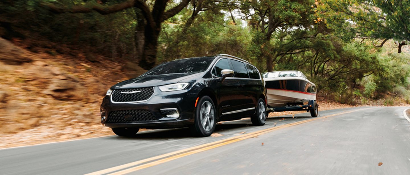 Imagen Una Chrysler Pacifica Pinnacle 2025 negra circulando por una carretera, remolcando una lancha. El fondo está borroso para indicar que el vehículo está en movimiento.