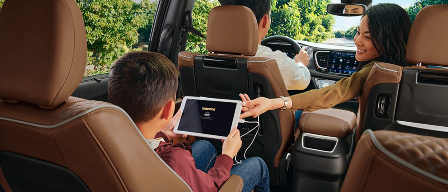 Imagen El interior de la Chrysler Pacifica Pinnacle 2025 con un hombre conduciendo, una mujer estirando la mano hacia la parte trasera y un niño en la segunda fila sujetando una tablet conectada a un tomacorriente.
