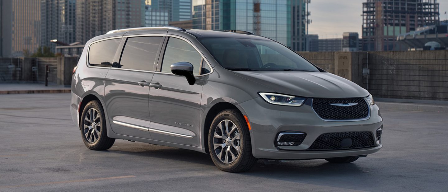 Se muestra el ángulo delantero del lado del pasajero de una Chrysler Pacifica Pinnacle Plug-in Hybrid 2025 gris estacionada en el techo de una estructura de estacionamiento, con edificios altos de fondo.