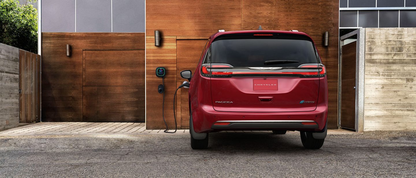Se muestra un ángulo trasero de una Chrysler Pacifica Pinnacle Hybrid enchufable 2025 roja estacionada afuera de un garaje, con un cable de carga enchufado en su puerto de carga.