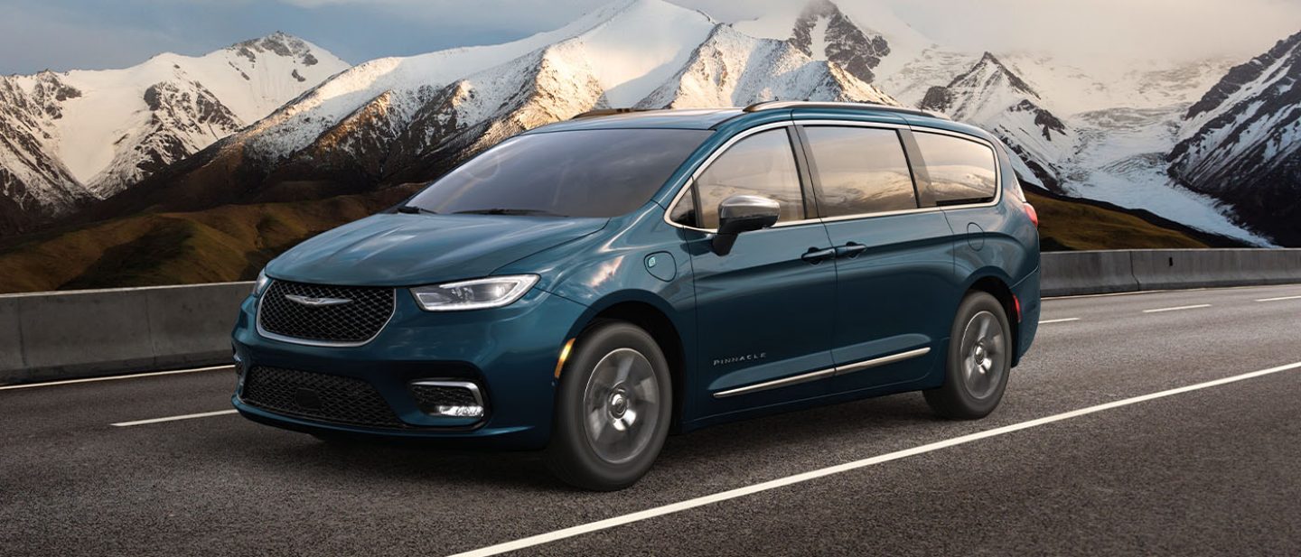 Se muestra una Chrysler Pacifica Pinnacle Plug-In Hybrid 2025 azul circulando por una autopista, con montañas de fondo.