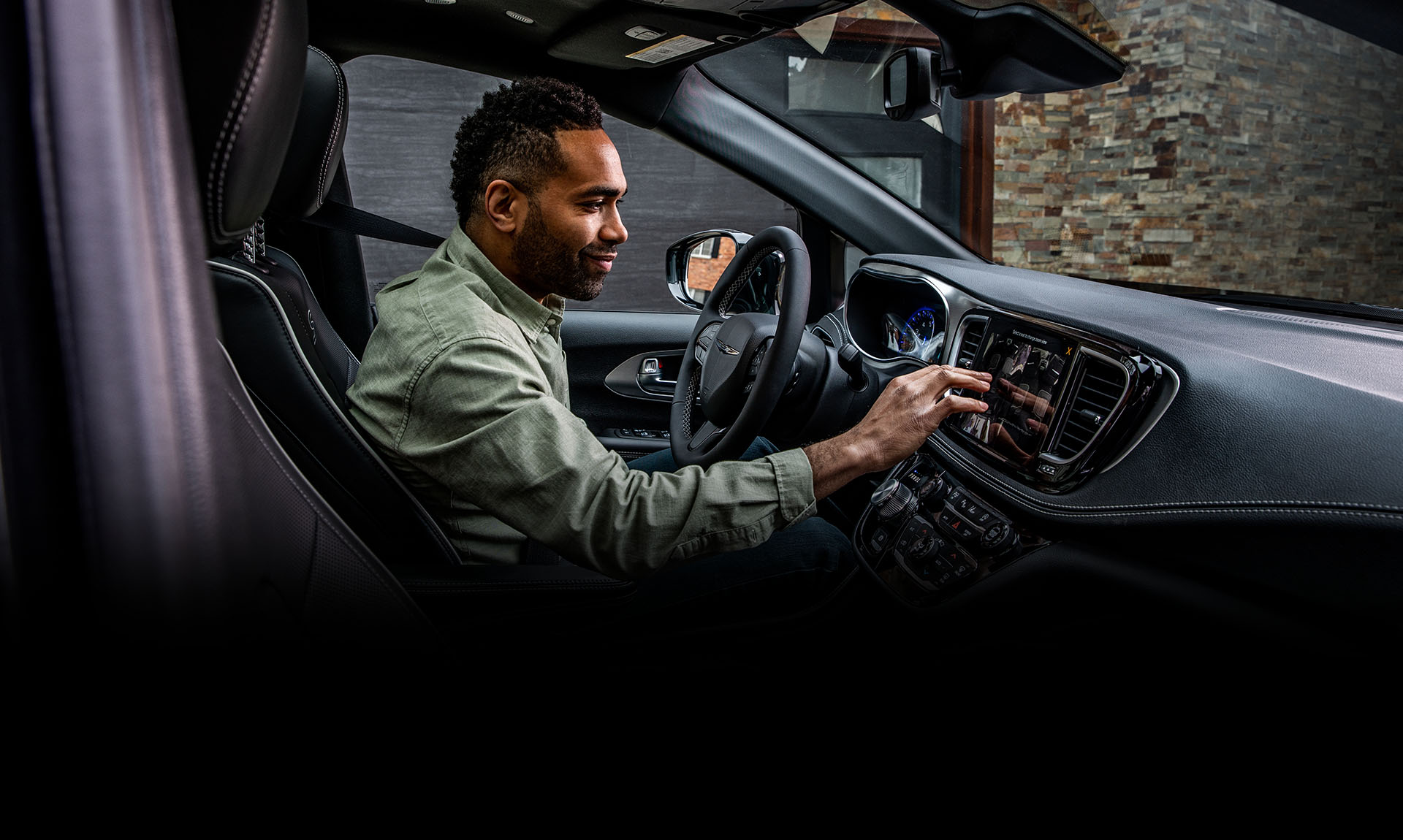 El área de los asientos delanteros en la Chrysler Pacifica Hybrid 2025 enchufable con el conductor haciendo una selección en la pantalla táctil Uconnect.