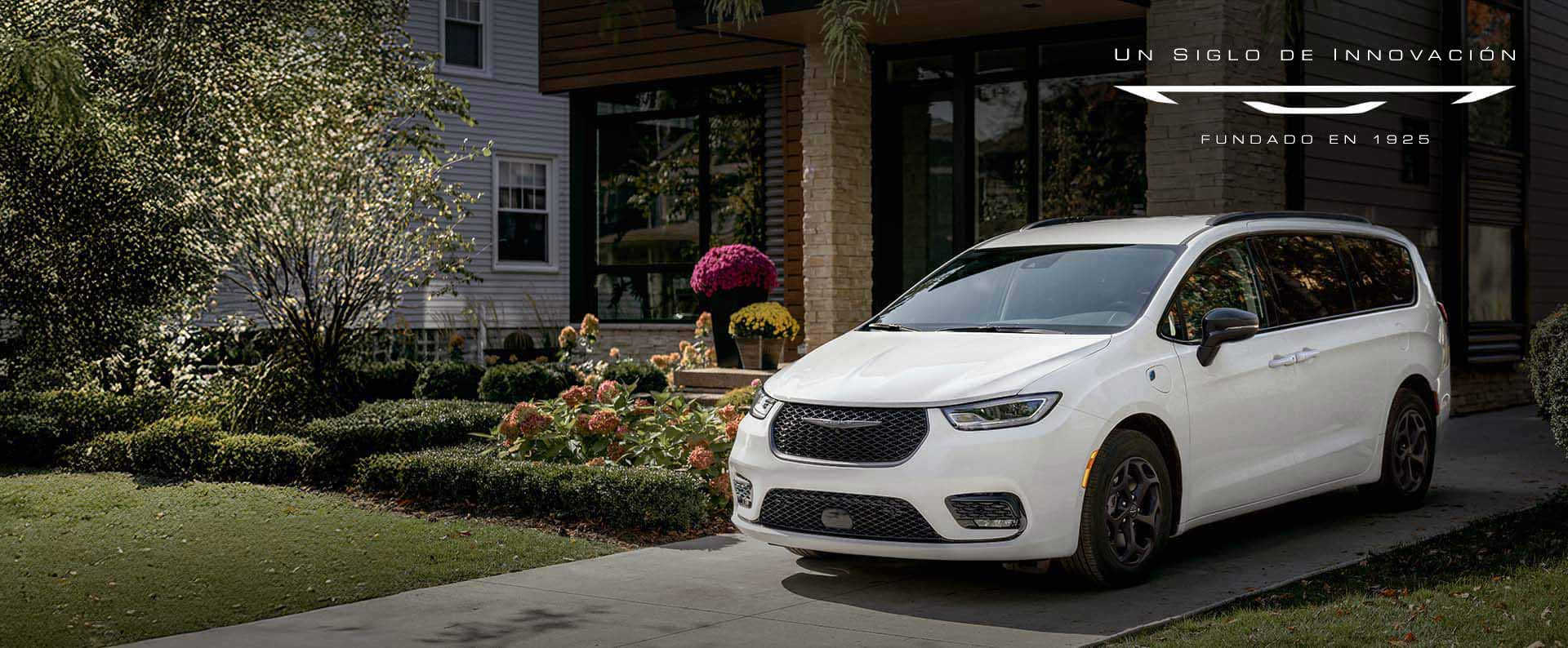 Ángulo delantero del lado del conductor de una Chrysler Pacifica Select Plug-in Hybrid 2025 blanca con el paquete apariencia Premium S, estacionada en la entrada de una casa en un vecindario. Un siglo de innovación.
