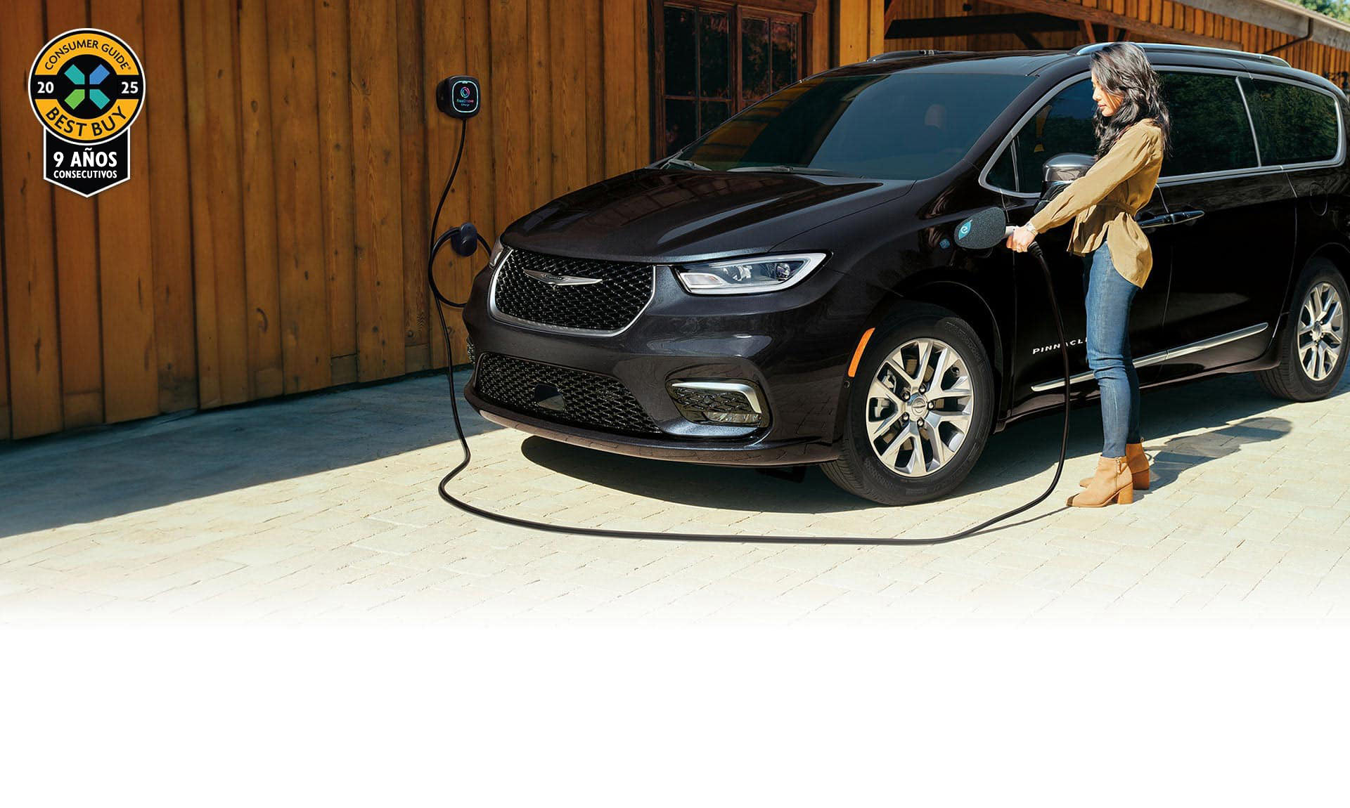 Ángulo delantero del lado del conductor de una Chrysler Pacifica Pinnacle Hybrid 2025 enchufable de color negro, estacionada fuera de un edificio con una estación de carga. Una mujer enchufando el cable de carga de la estación en el puerto de carga del vehículo. Premio Consumer Guide Best Buy 2025.