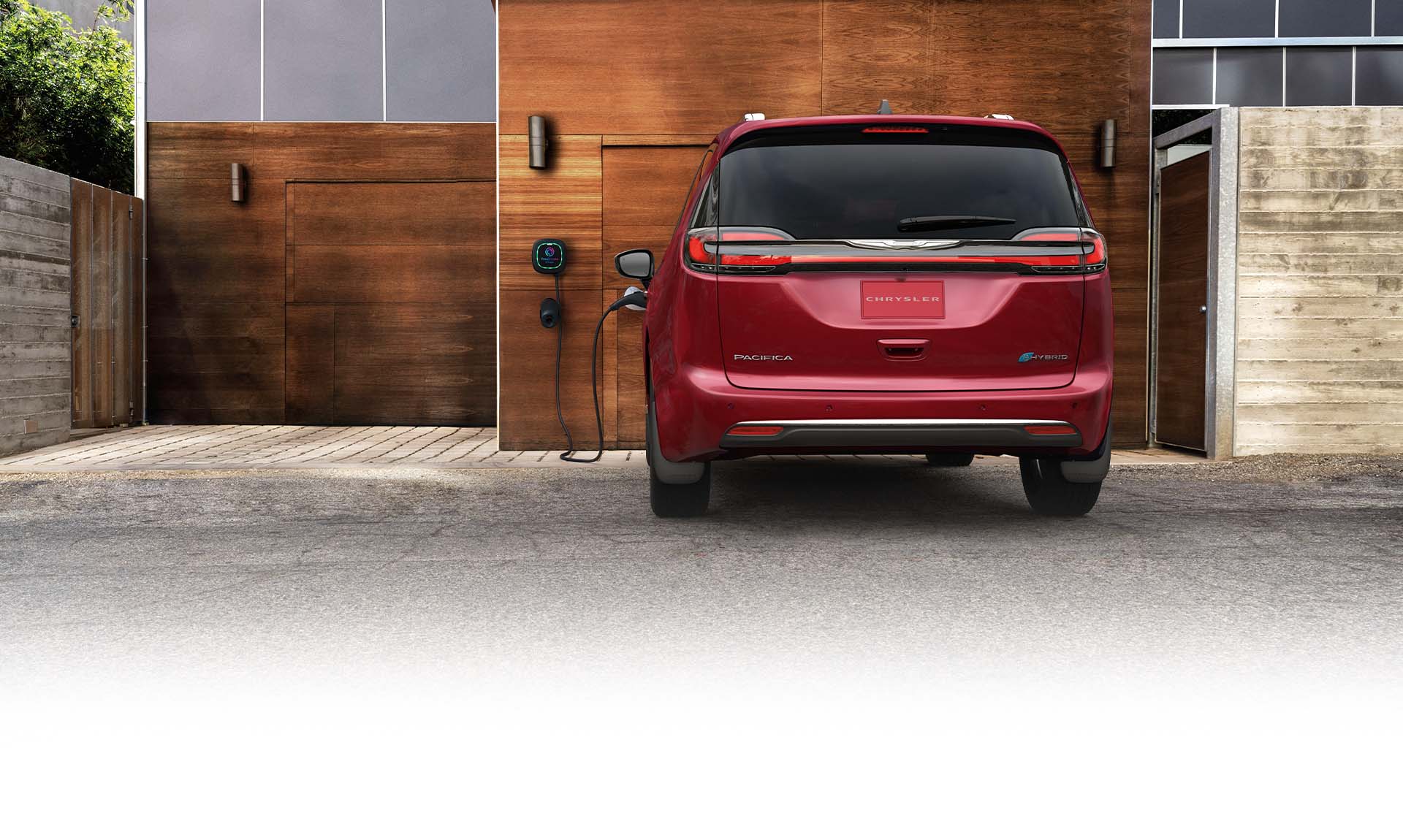 Ángulo trasero de una Chrysler Pacifica Pinnacle Plug-In Hybrid 2025 roja estacionada fuera de un garaje con una estación de carga de nivel 2 integrada, con un cable de carga enchufado en el puerto de carga del vehículo.