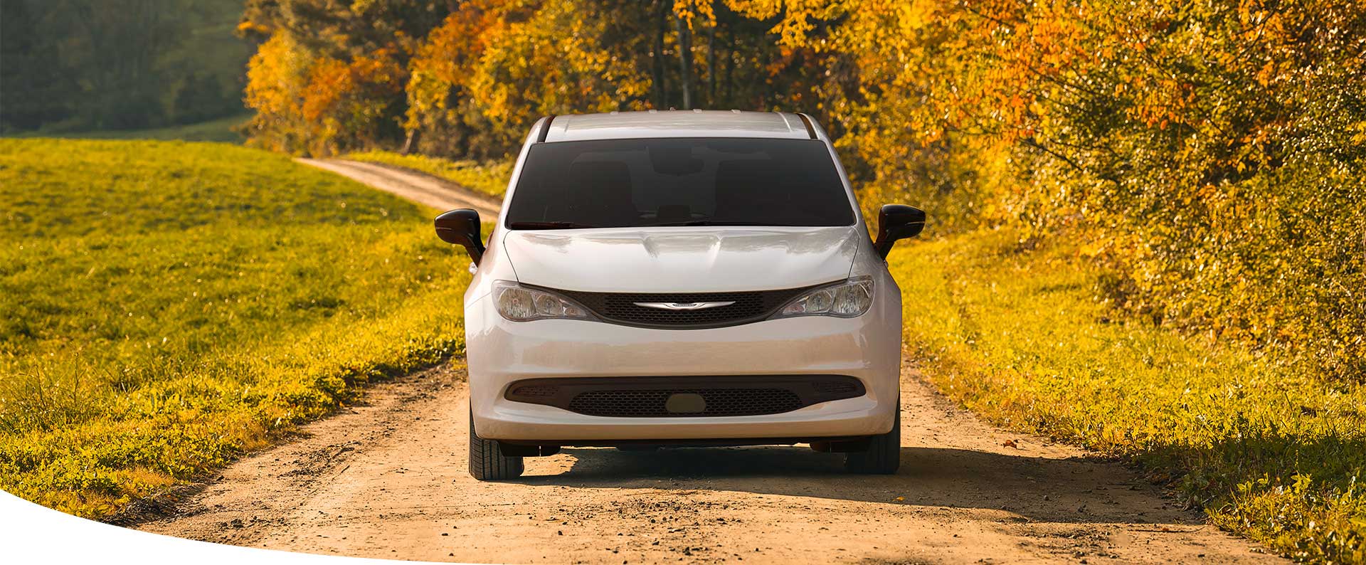 Un ángulo frontal de una Chrysler Voyager LX 2025 blanca andando por un camino de tierra en el campo.