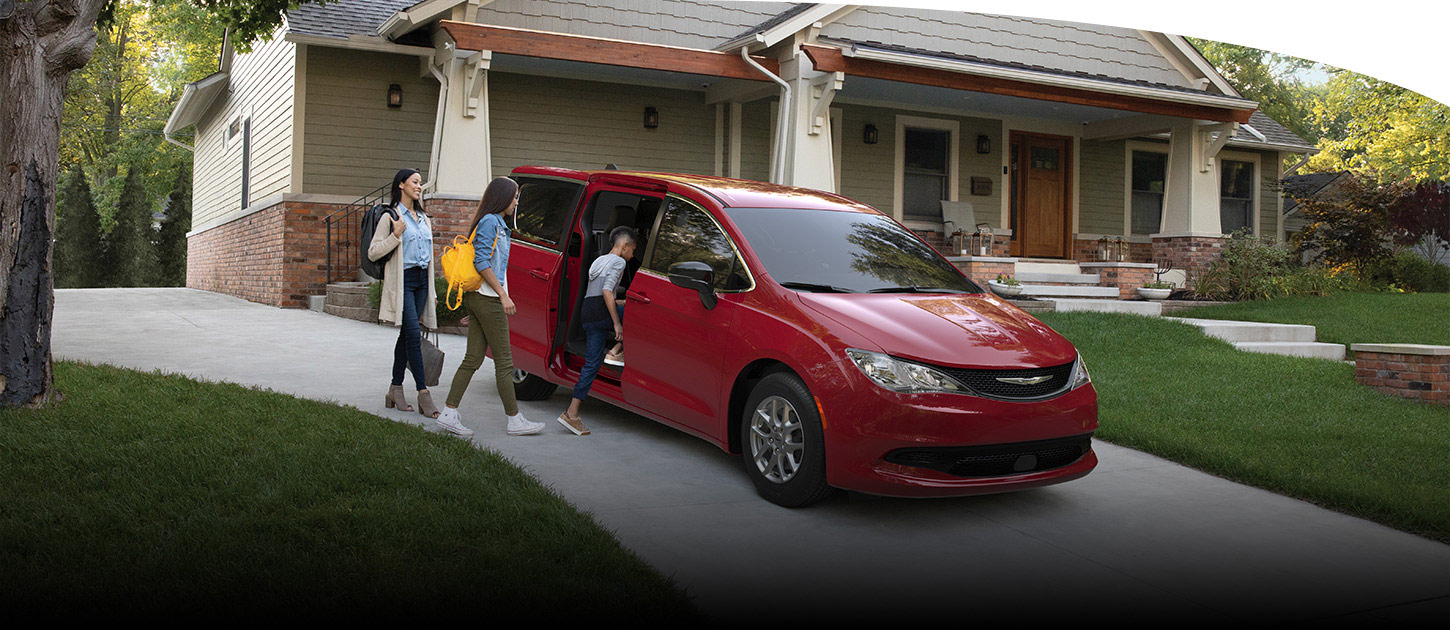 Una Chrysler Voyager LX 2025 roja estacionada en la entrada de una casa con la puerta lateral deslizable abierta. Un niño sube al vehículo mientras una mujer lo acompaña.