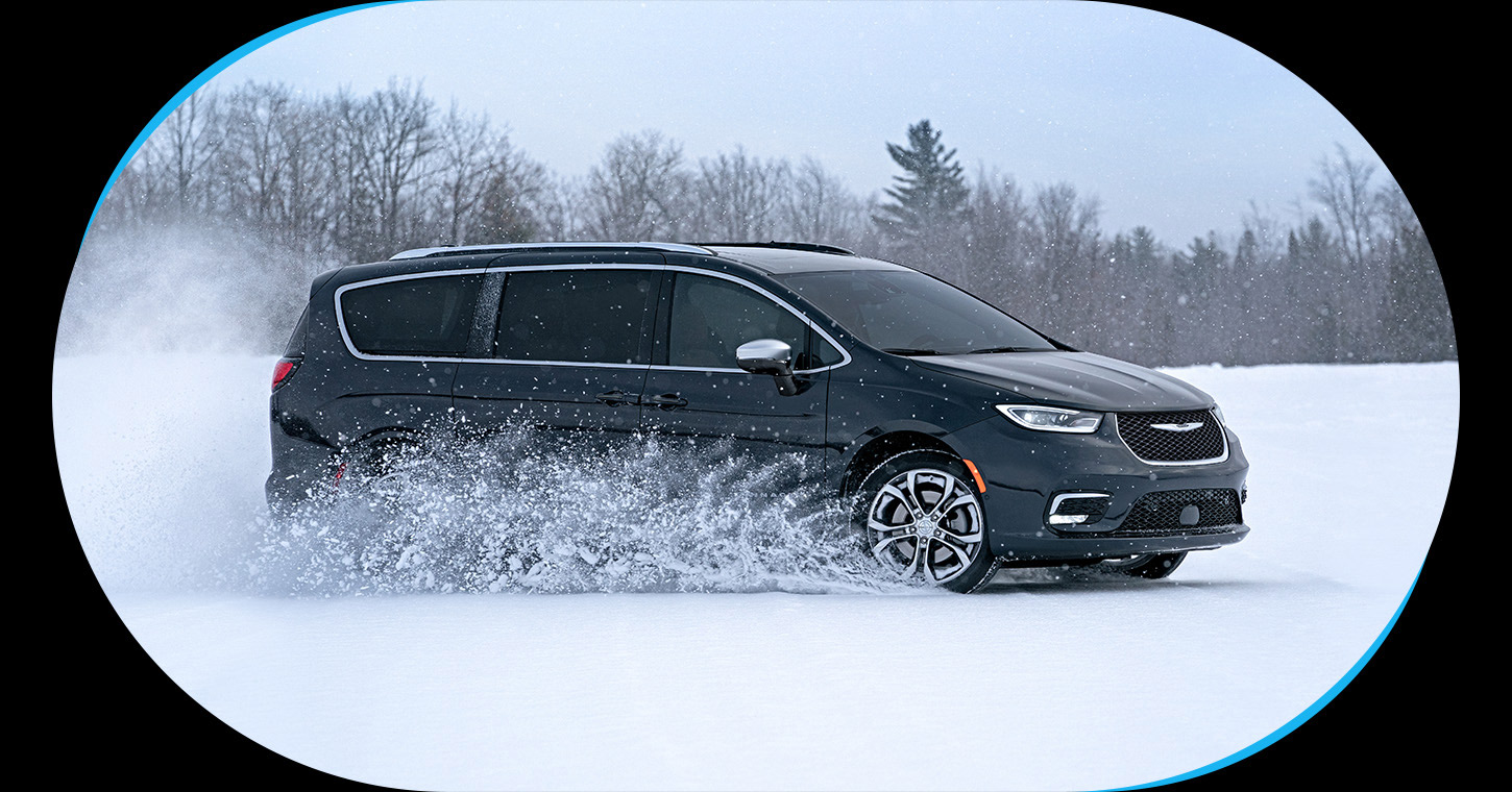 Perfil del lado del pasajero de una Chrysler Pacifica Pinnacle 2026 negra circulando por un camino cubierto de nieve en el bosque.