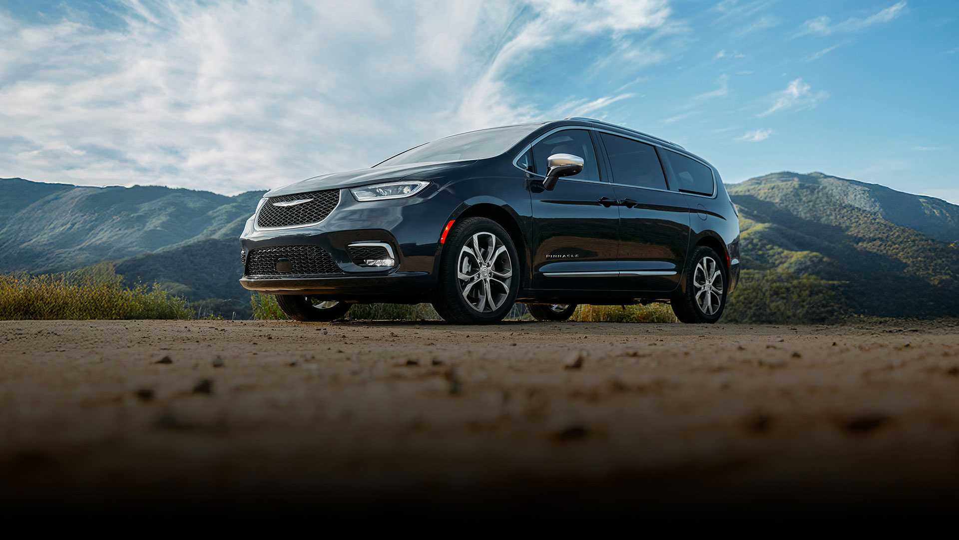 Ángulo delantero del lado del conductor de una Chrysler Pacifica Pinnacle 2026 negra estacionada en un claro con montañas de fondo.
