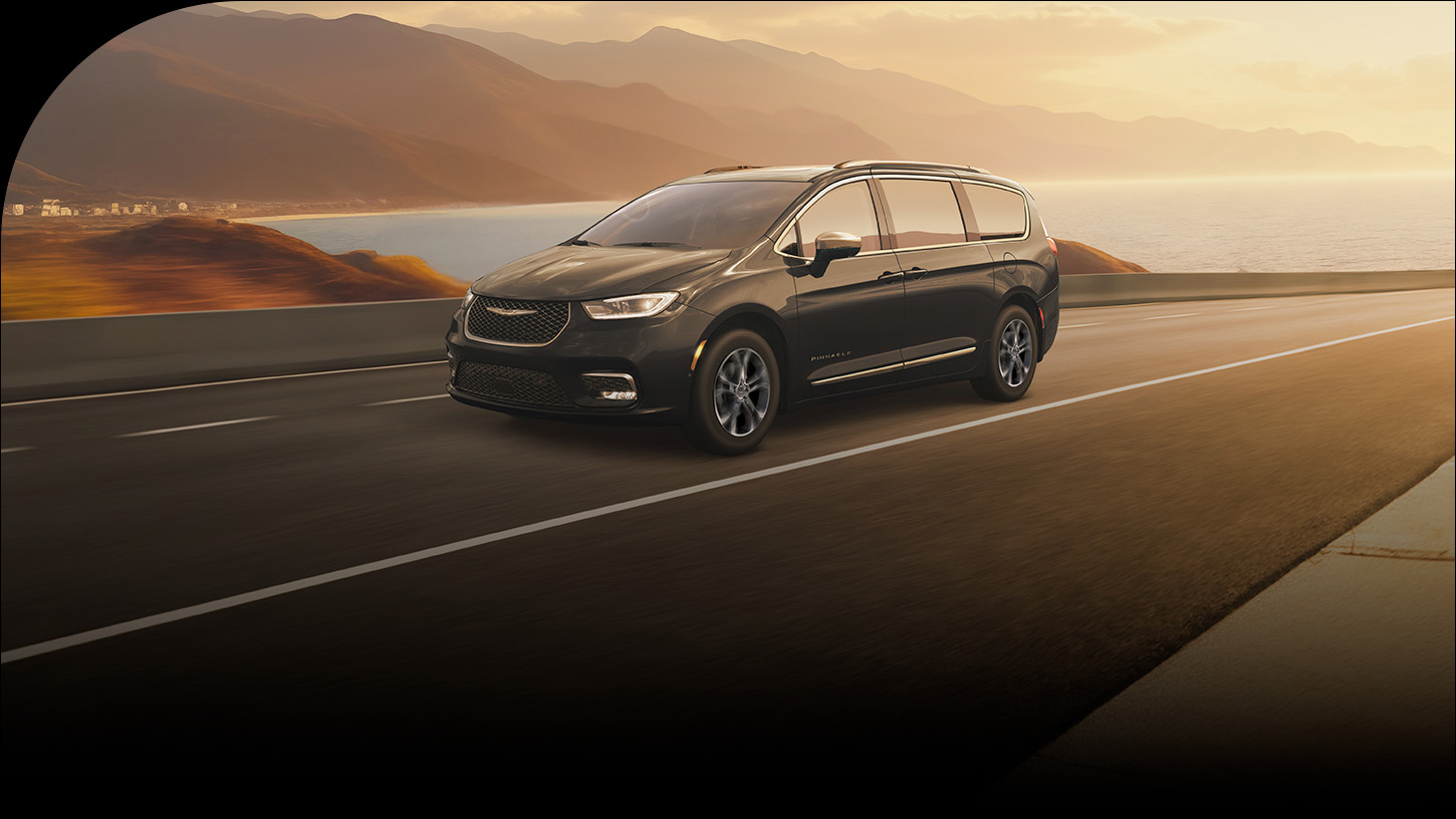 Una Chrysler Pacifica Pinnacle 2026 negra recorriendo una carretera al atardecer, con un lago y montañas de fondo.