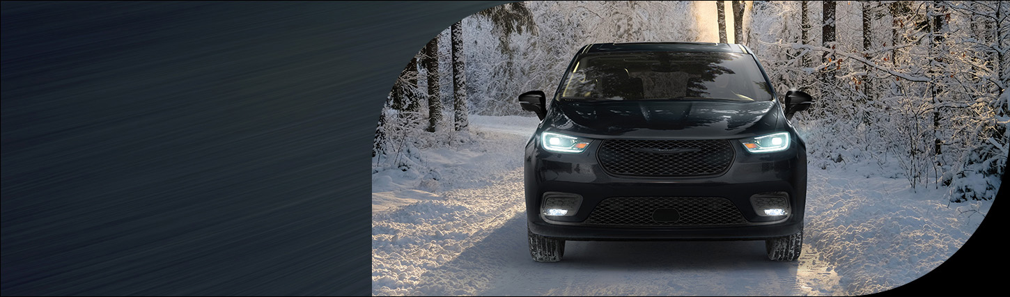 Ángulo frontal de una Chrysler Pacifica Limited 2026 negra, con los faros delanteros encendidos, recorriendo un camino nevado en el bosque.