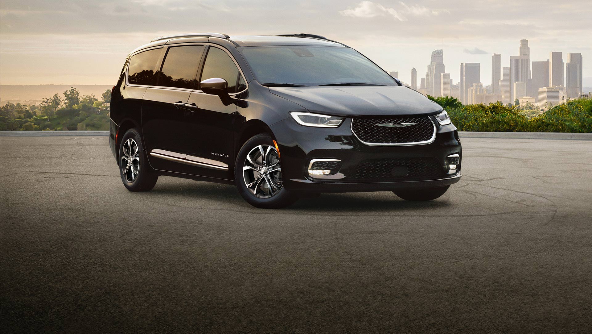 Ángulo delantero del lado del pasajero de una Chrysler Pacifica Pinnacle 2026 negra estacionada con un paisaje urbano de fondo.