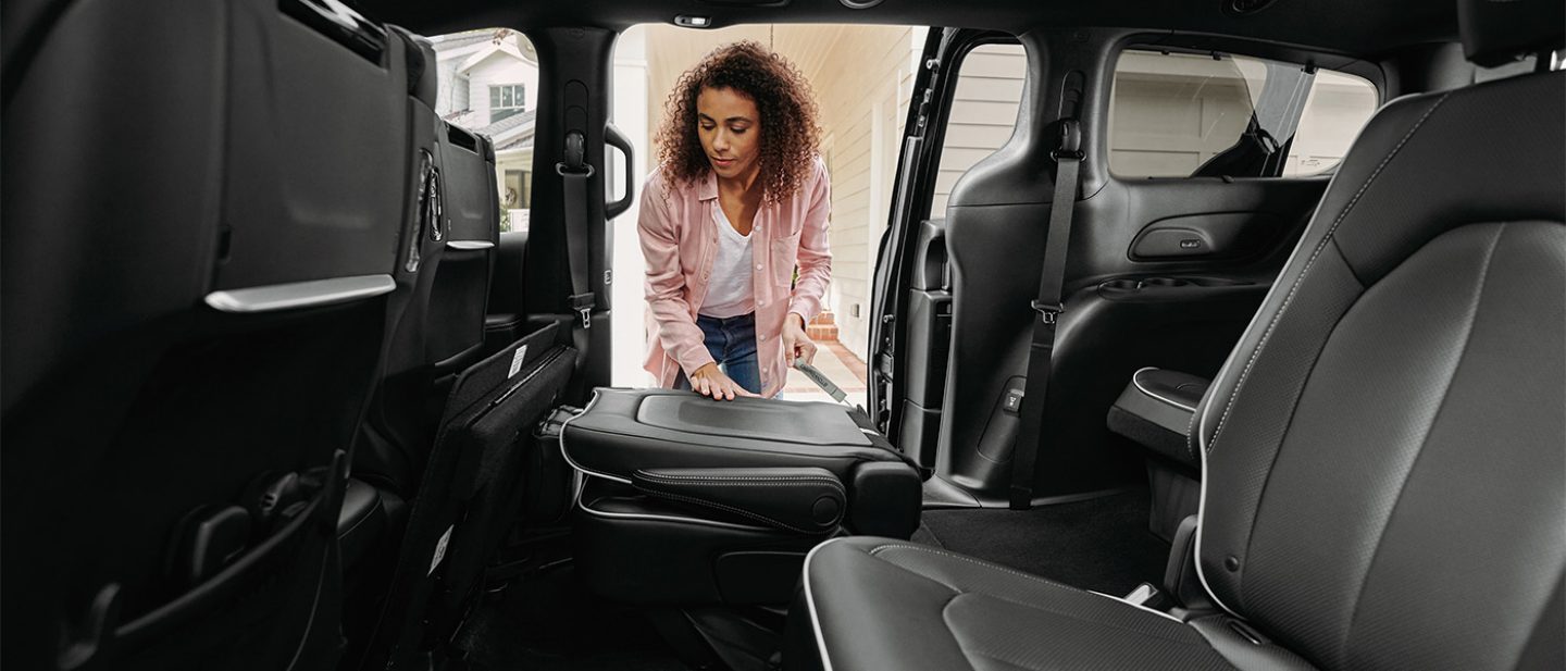Se muestra el interior de la Chrysler Pacifica 2026 con una mujer junto a la puerta corrediza del lado del pasajero abierta. Está plegando uno de los asientos de la segunda fila hasta el piso.