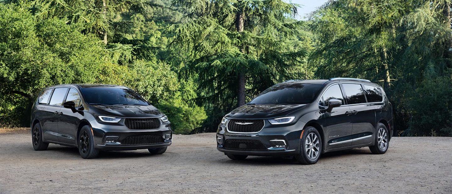 Se muestran dos modelos Chrysler Pacifica Plug-in Hybrid 2026 de color azul estacionados perpendicularmente uno al otro en un parque. A la izquierda, una Pacifica Plug-in Hybrid Select con el paquete de apariencia Premium S, y a la derecha, una Pacifica Plug-in Hybrid Pinnacle.