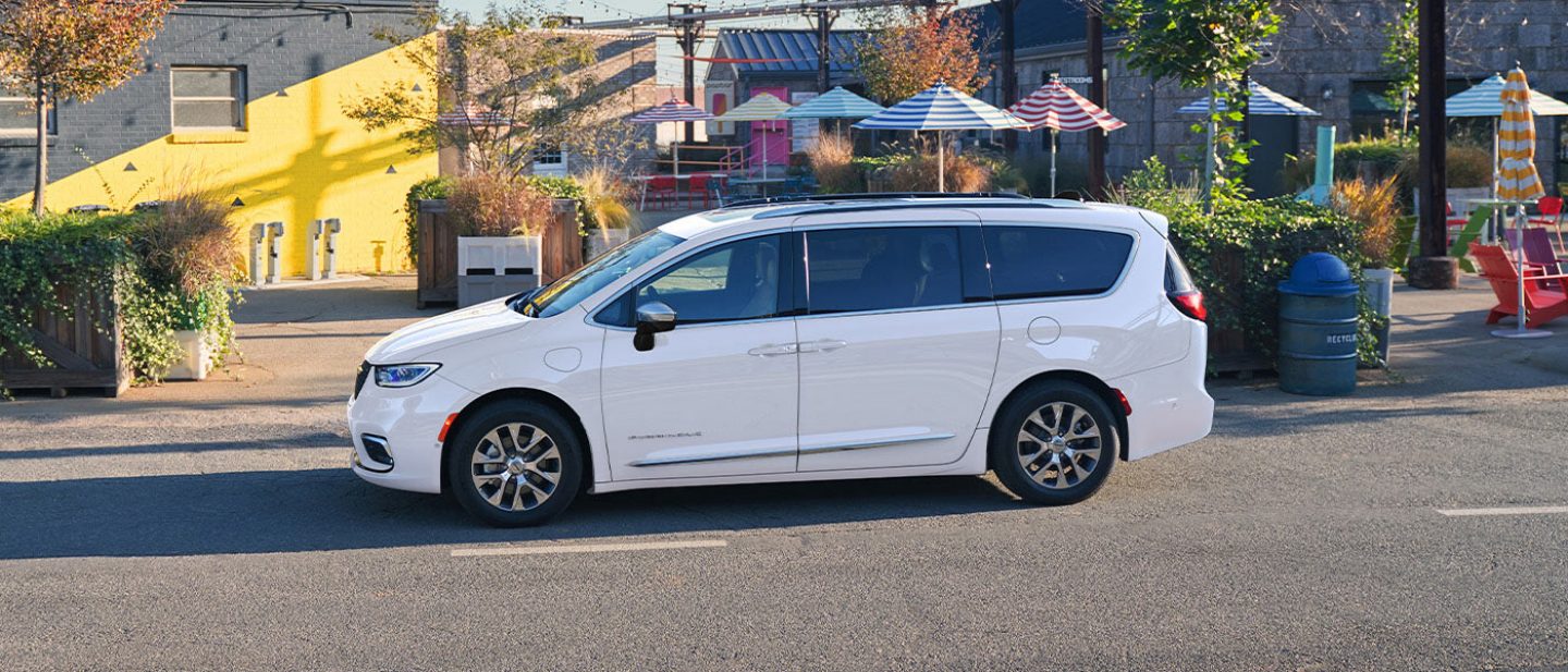 Se muestra el perfil del lado del conductor de una Chrysler Pacifica Plug-in Hybrid Pinnacle 2026 de color blanco estacionada en un lugar turístico.