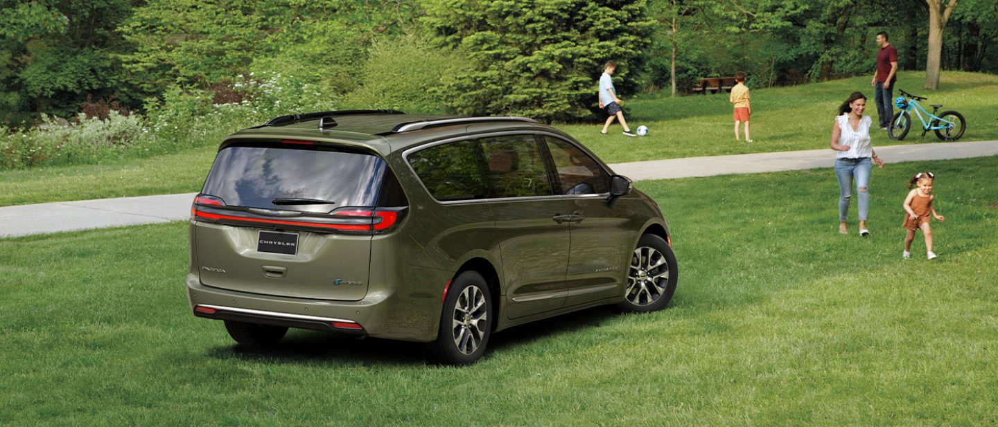 Se muestra un ángulo trasero del lado del pasajero de una Chrysler Pacifica Plug-in Hybrid 2026 estacionada en el césped de un parque con niños y adultos cerca.