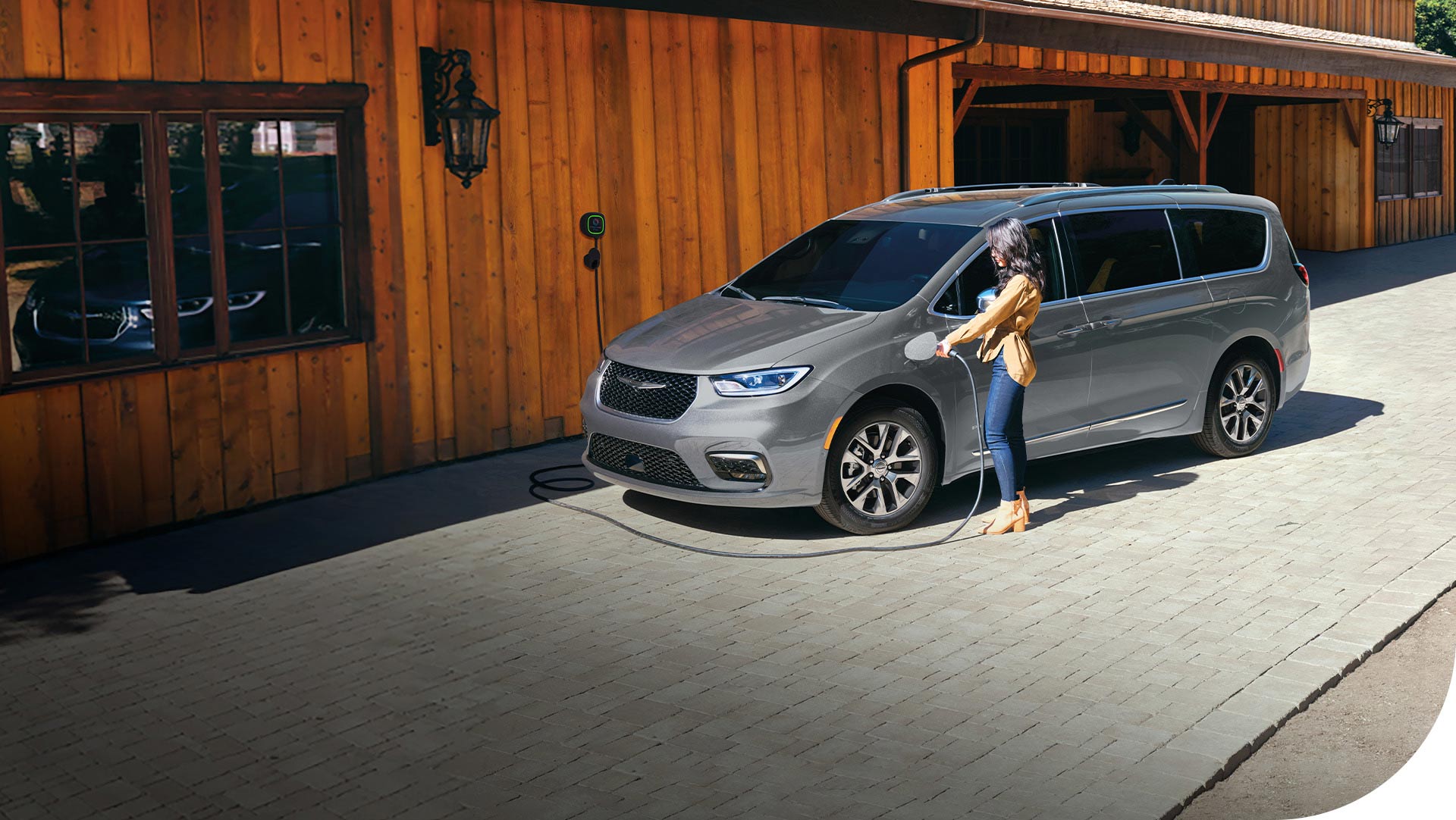 Ángulo delantero del lado del conductor de una Chrysler Pacifica Plug-In Hybrid Pinnacle 2026 de color plateado estacionada en un mercado agrícola que cuenta con una estación de carga integrada, con una mujer enchufando el cable de carga en el puerto de carga de la minivan.