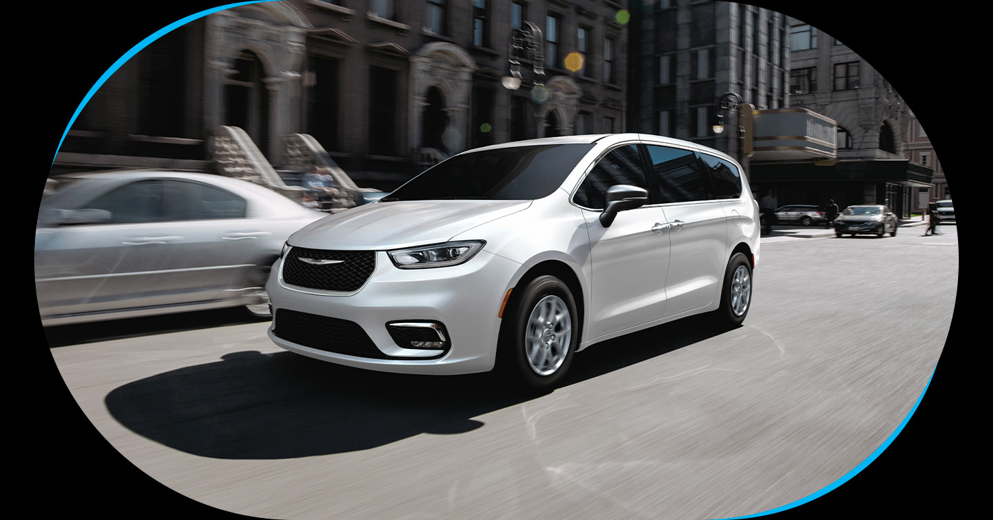 Una Chrysler Pacifica Select 2026 blanca circulando por una calle urbana ajetreada.