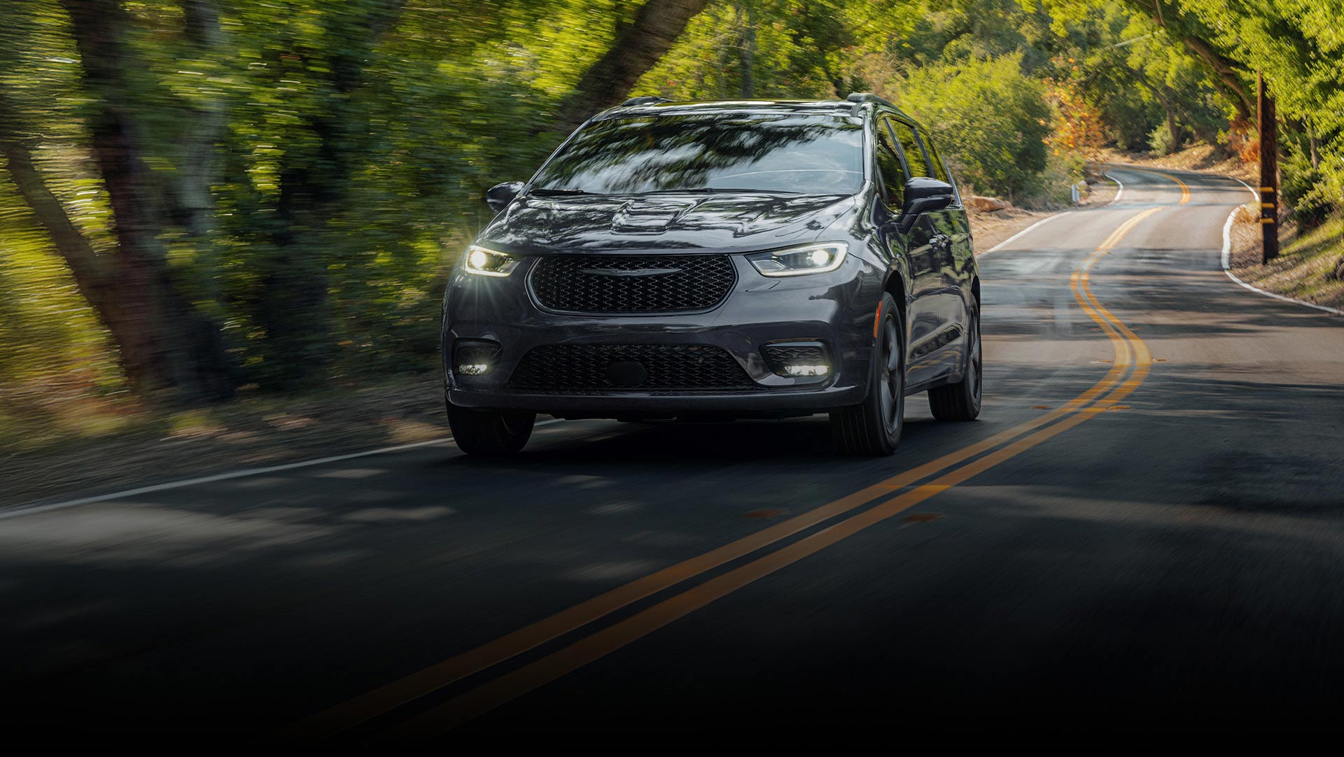 Una Chrysler Pacifica Limited 2026 gris circulando por una carretera sinuosa en el bosque.