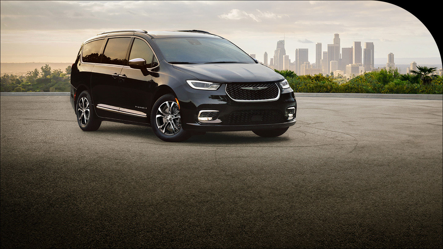 Ángulo delantero del lado del pasajero de una Chrysler Pacifica Pinnacle 2026 negra estacionada con un paisaje urbano de fondo.