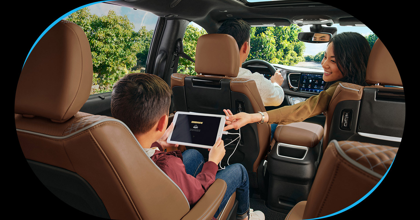 El interior de la Chrysler Pacifica Pinnacle 2026 con un hombre conduciendo, una mujer estirando la mano hacia la parte trasera y un niño en la segunda fila sujetando una tablet conectada a un tomacorriente.