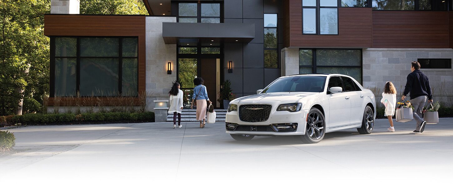 Un Chrysler 300 300S blanco 2023 estacionado en el camino de entrada de una amplia casa contemporánea, con una familia de cuatro personas caminando desde el vehículo hacia la puerta principal.