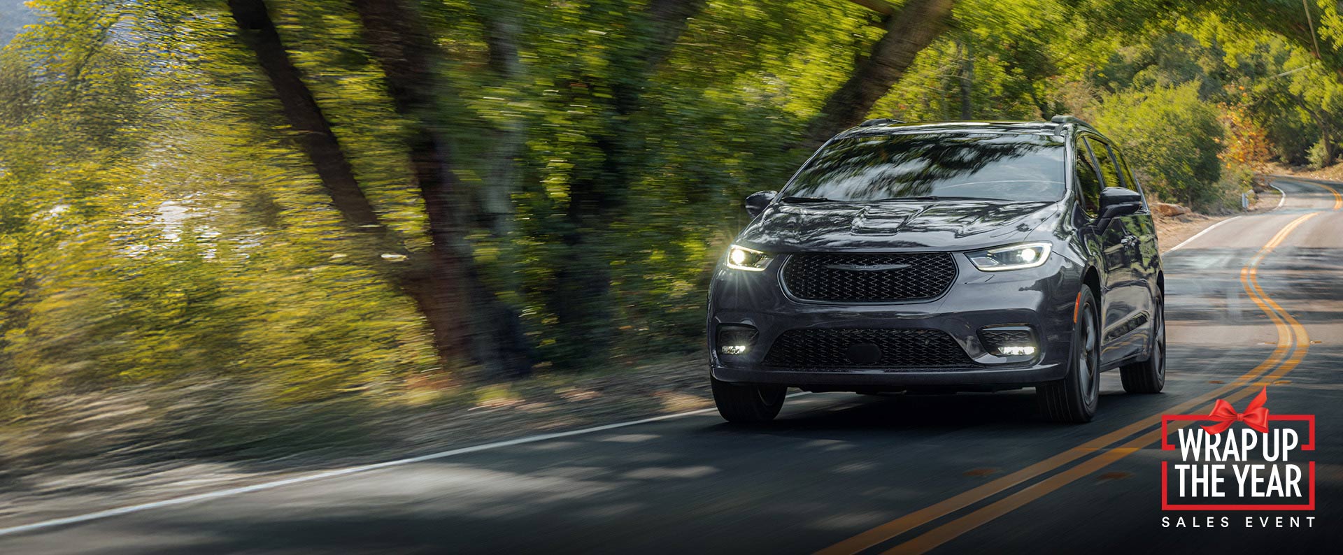 Una Chrysler Pacifica Limited 2026 gris circulando por una carretera sinuosa en el bosque. Evento de ventas Fin de año.