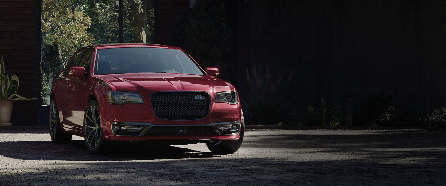 Vista frontal de medio perfil de un Chrysler 300C 2023 rojo estacionado en la entrada de garaje de una casa moderna.