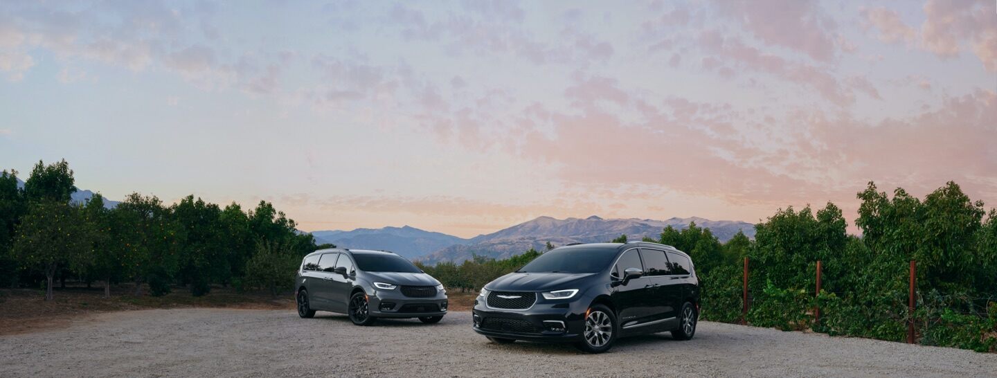 Dos modelos Chrysler Pacifica 2021: Limited con el paquete de apariencia S y Pinnacle Hybrid, estacionados en un claro de gravilla entre árboles plantados en filas.